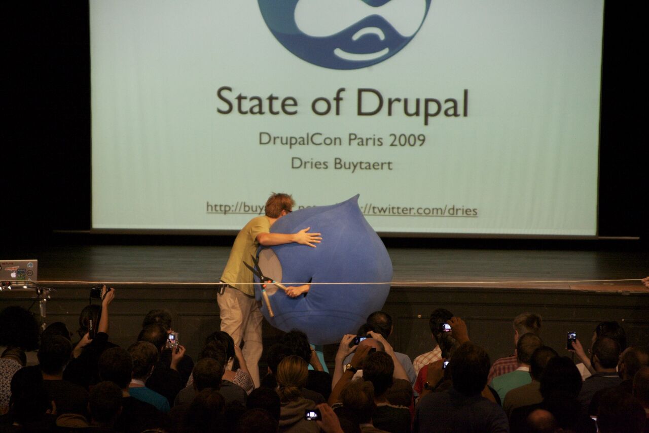 A speaker on stage hugs a large blue Drupal mascot holding large scissors while an audience takes photos.