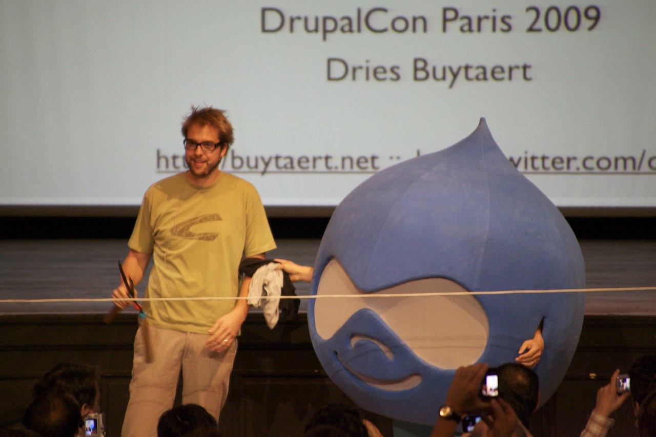 A man with scissors prepares to cut a ribbon alongside a person in a large blue Drupal mascot costume.