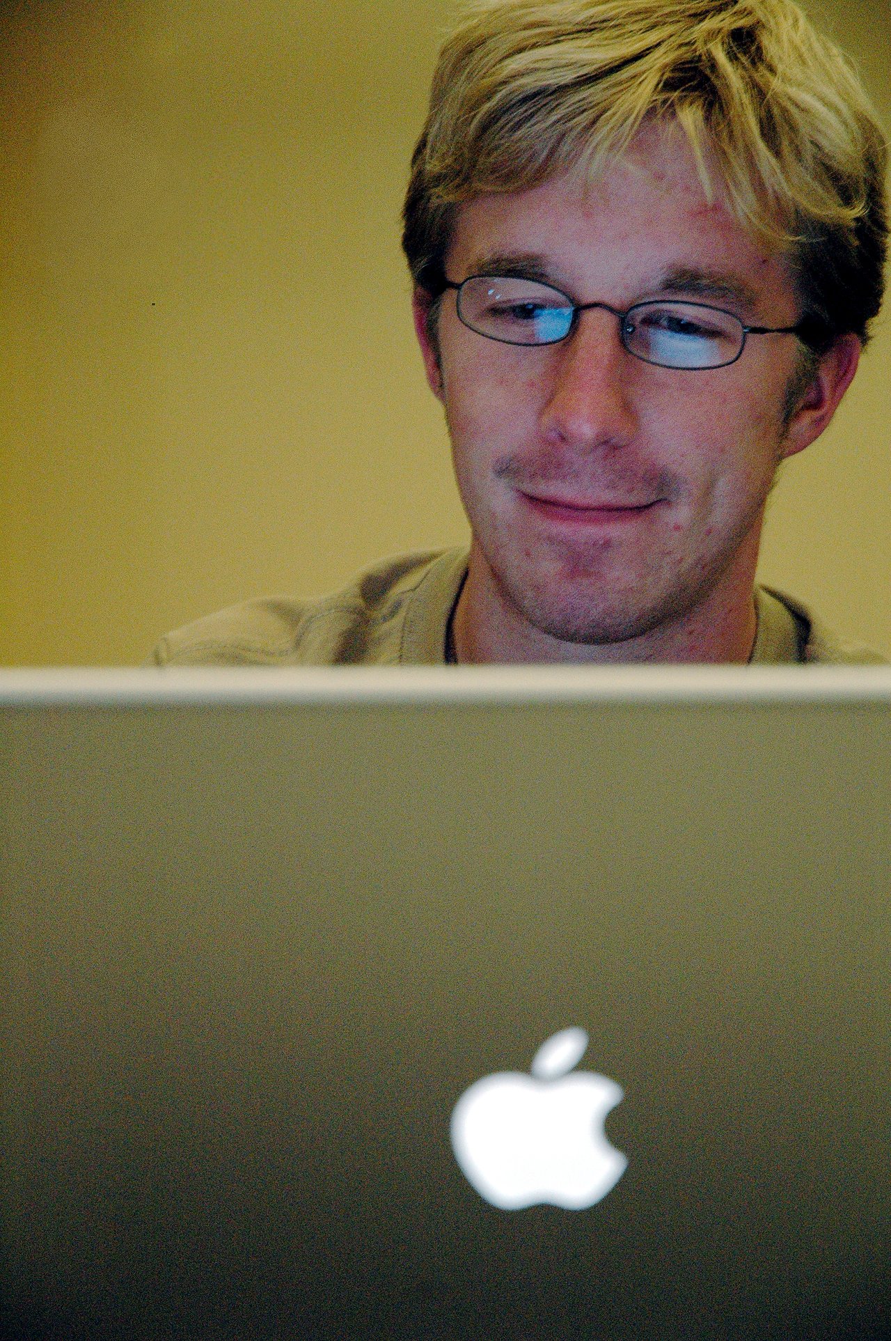 A person with glasses and blond hair is looking at a laptop screen with a focused expression.