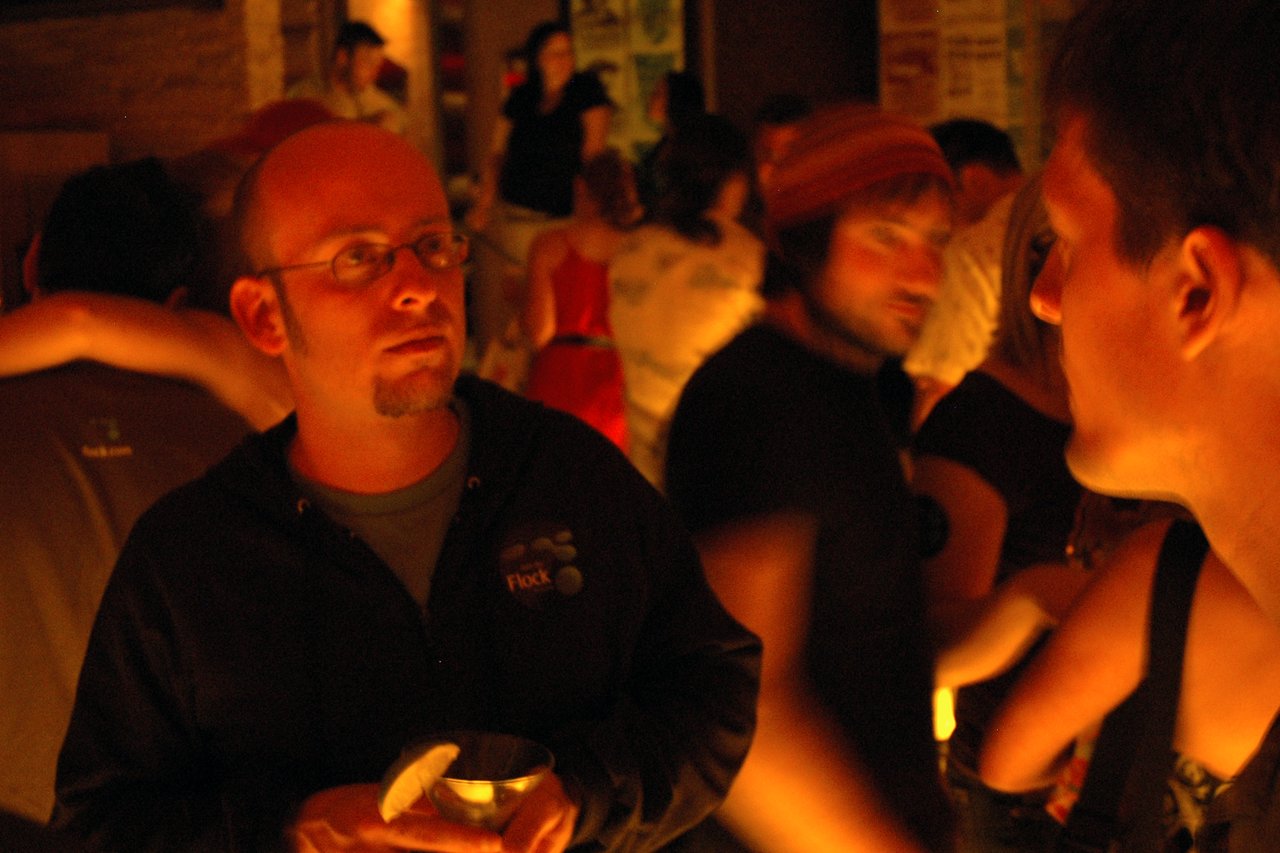 A man in glasses and a hoodie holds a drink while talking to another person in a dimly lit setting.