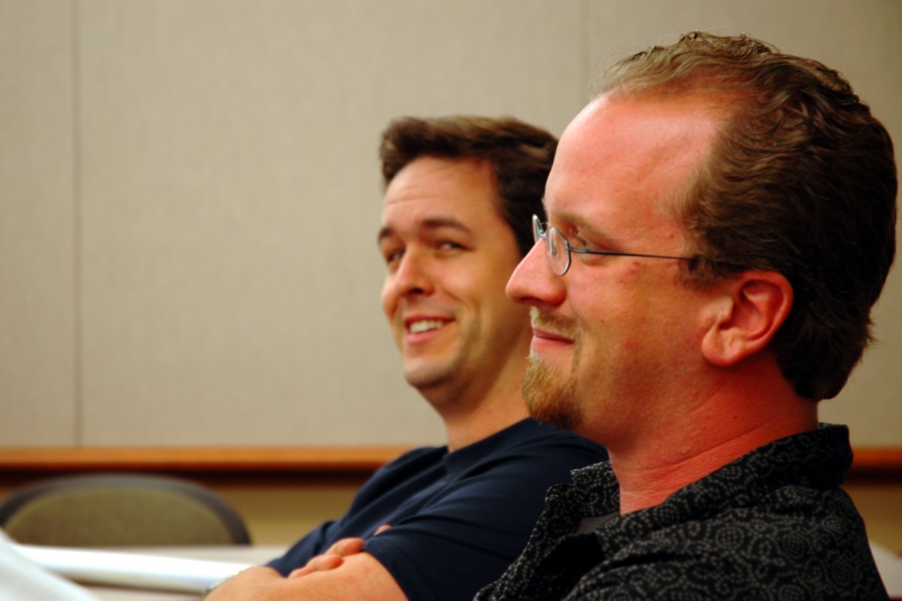 Two men sitting in a conference room, smiling and listening to a presentation at DrupalCon Portland 2005.