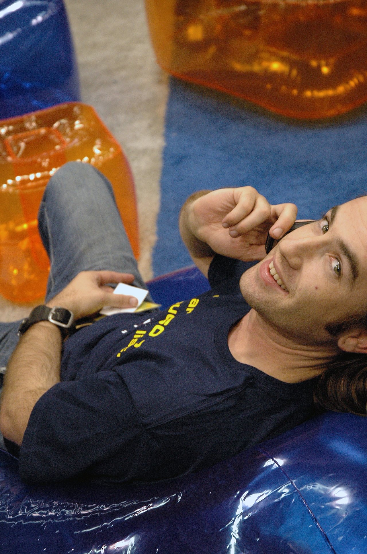 A man reclines on a blue inflatable chair, smiling while talking on a mobile phone and holding business cards.