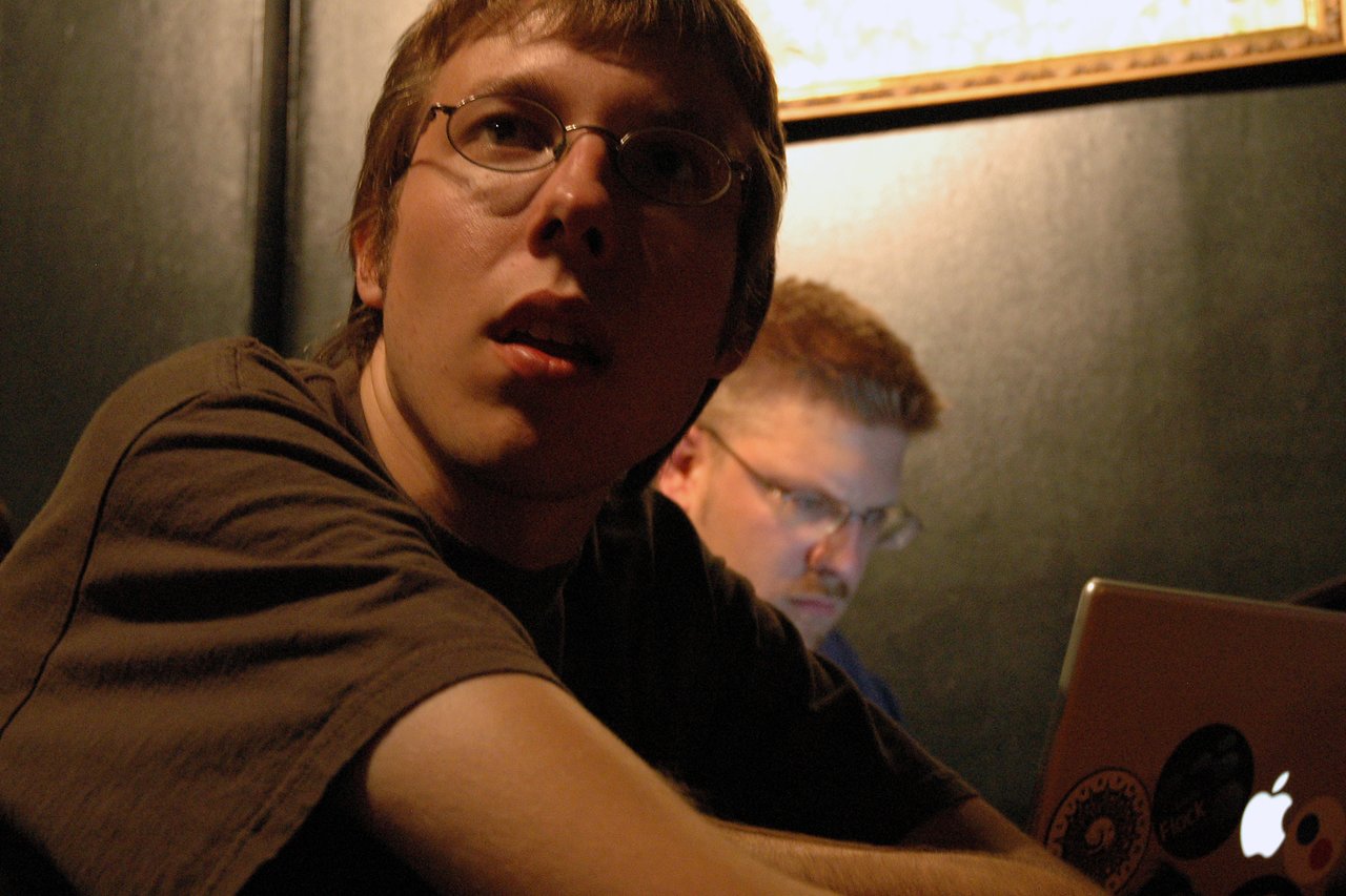 A person in glasses looks up with an attentive expression, while another person focuses on a laptop in the background.