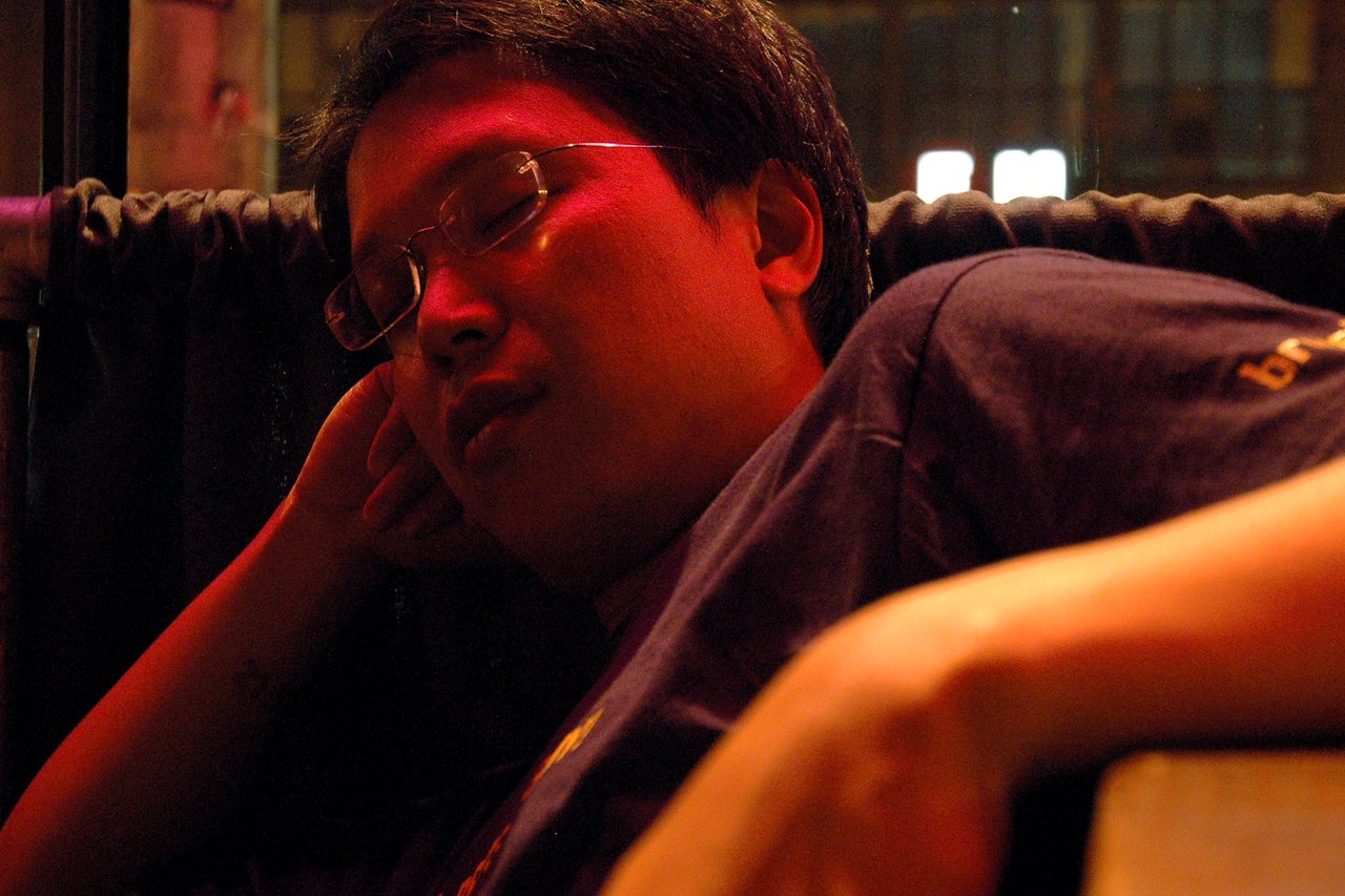 A person wearing glasses and a dark shirt is resting with their head tilted and eyes closed in dim lighting.