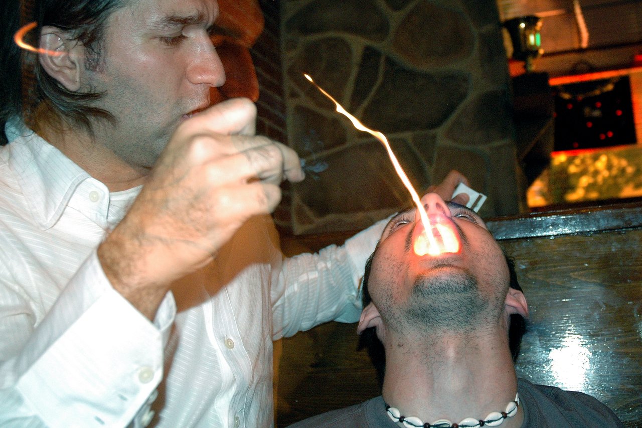A person leans back with fire coming from their mouth while another person in white holds a tool nearby.