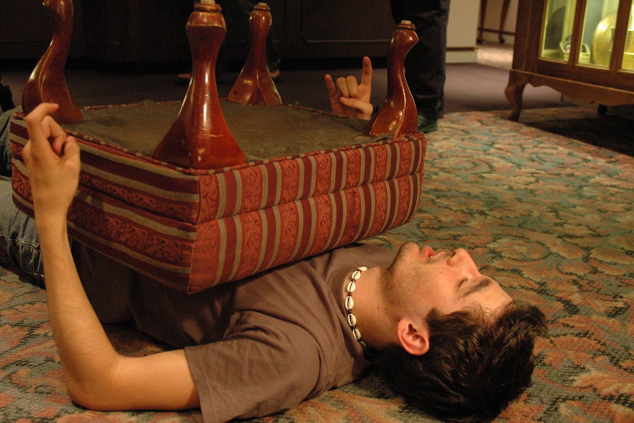 A person lies on the floor with an upside-down small table resting on their chest, making hand gestures.