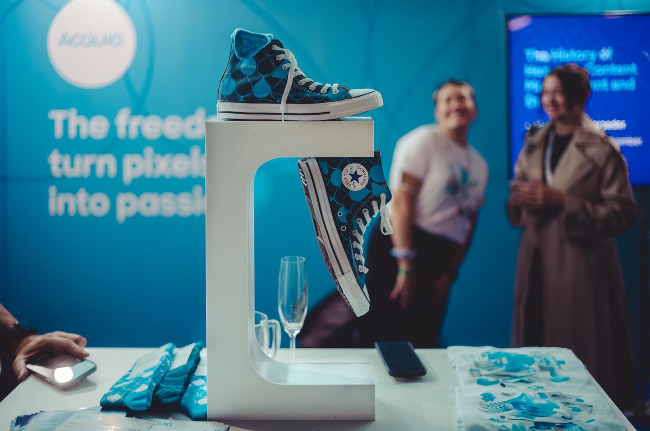 Blue Drupal-branded Converse shoes displayed at an Acquia booth, with people admiring them in the background.