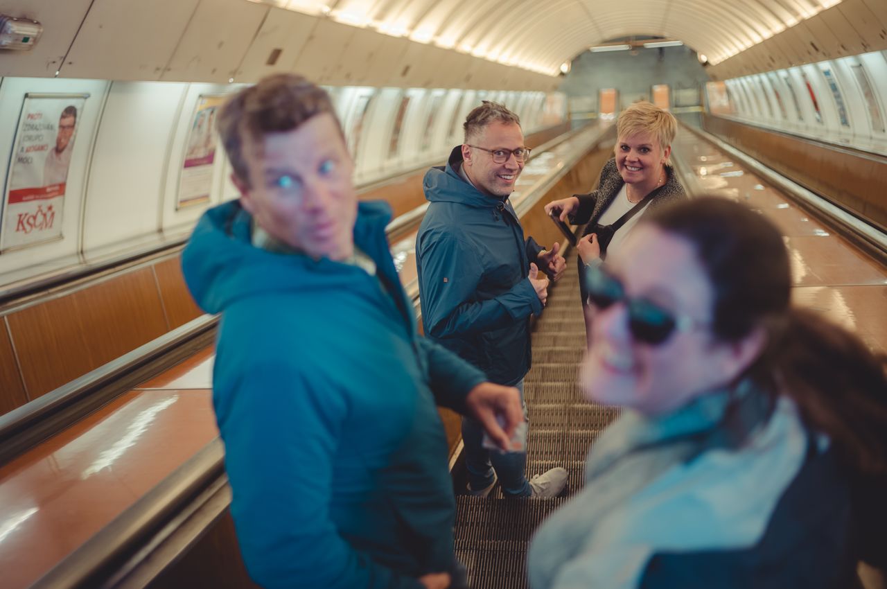 Four people ride a fast-moving escalator in a Prague metro station, smiling and looking back at the camera.