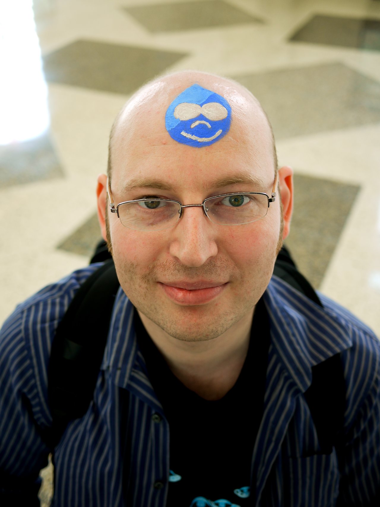 A person with glasses and a backpack has a blue Drupal logo painted on their forehead, smiling at the camera.