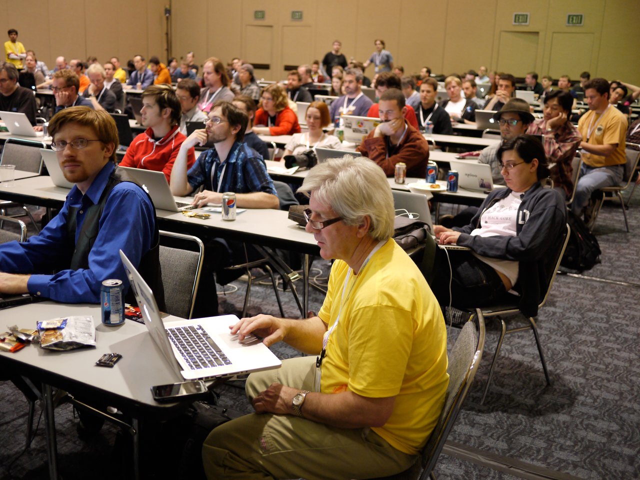 A group of developers attending a summit, working on laptops and listening to a presentation in a conference room.