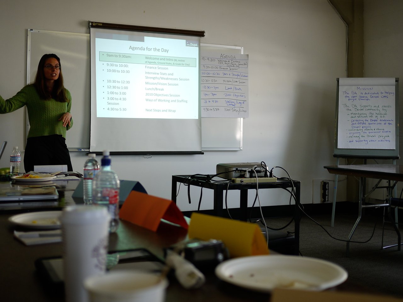 A woman stands at the front of a meeting room, gesturing while presenting an agenda on a projector screen.