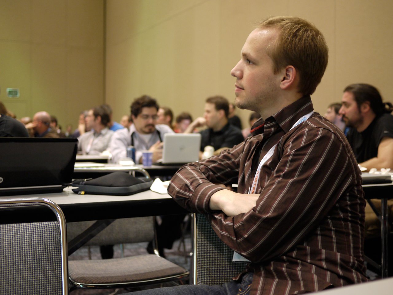 A man in a striped shirt sits at a conference table, attentively listening to a presentation.