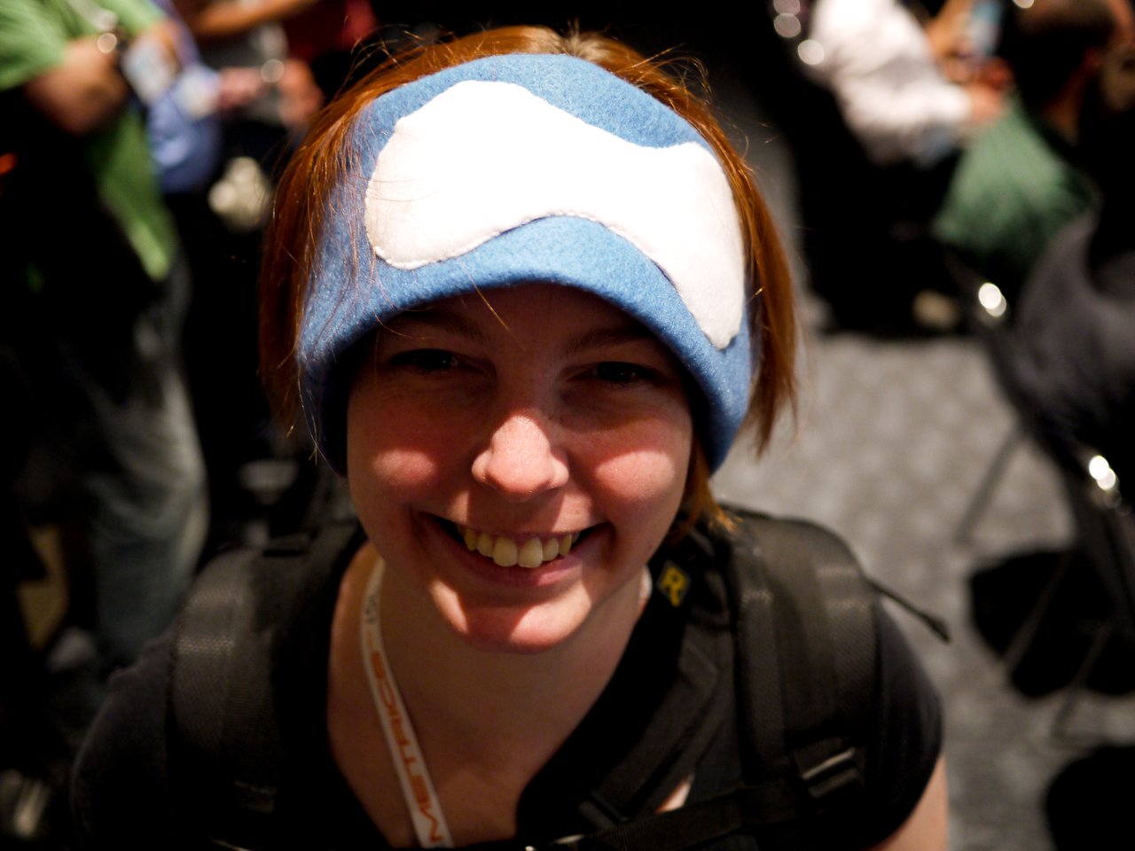 A person smiling while wearing a blue headband with a white design at a crowded event.