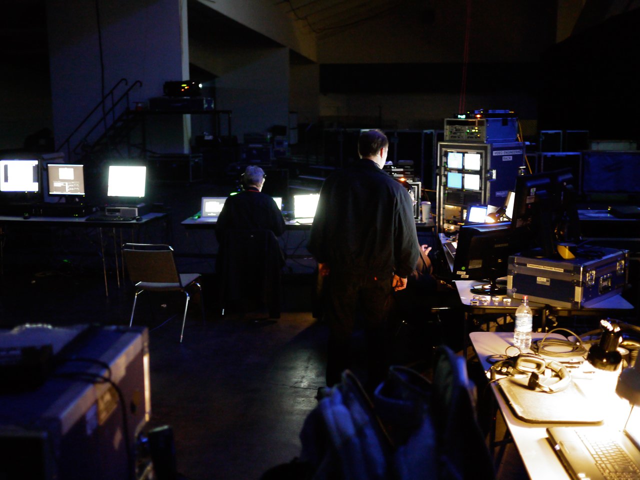 People working backstage at a keynote event, monitoring multiple screens and managing technical equipment in a dimly lit area.