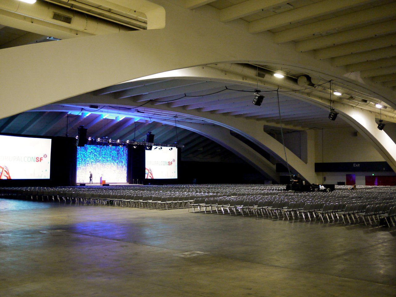 A large keynote hall with rows of empty chairs facing a stage, where a speaker stands near a podium.