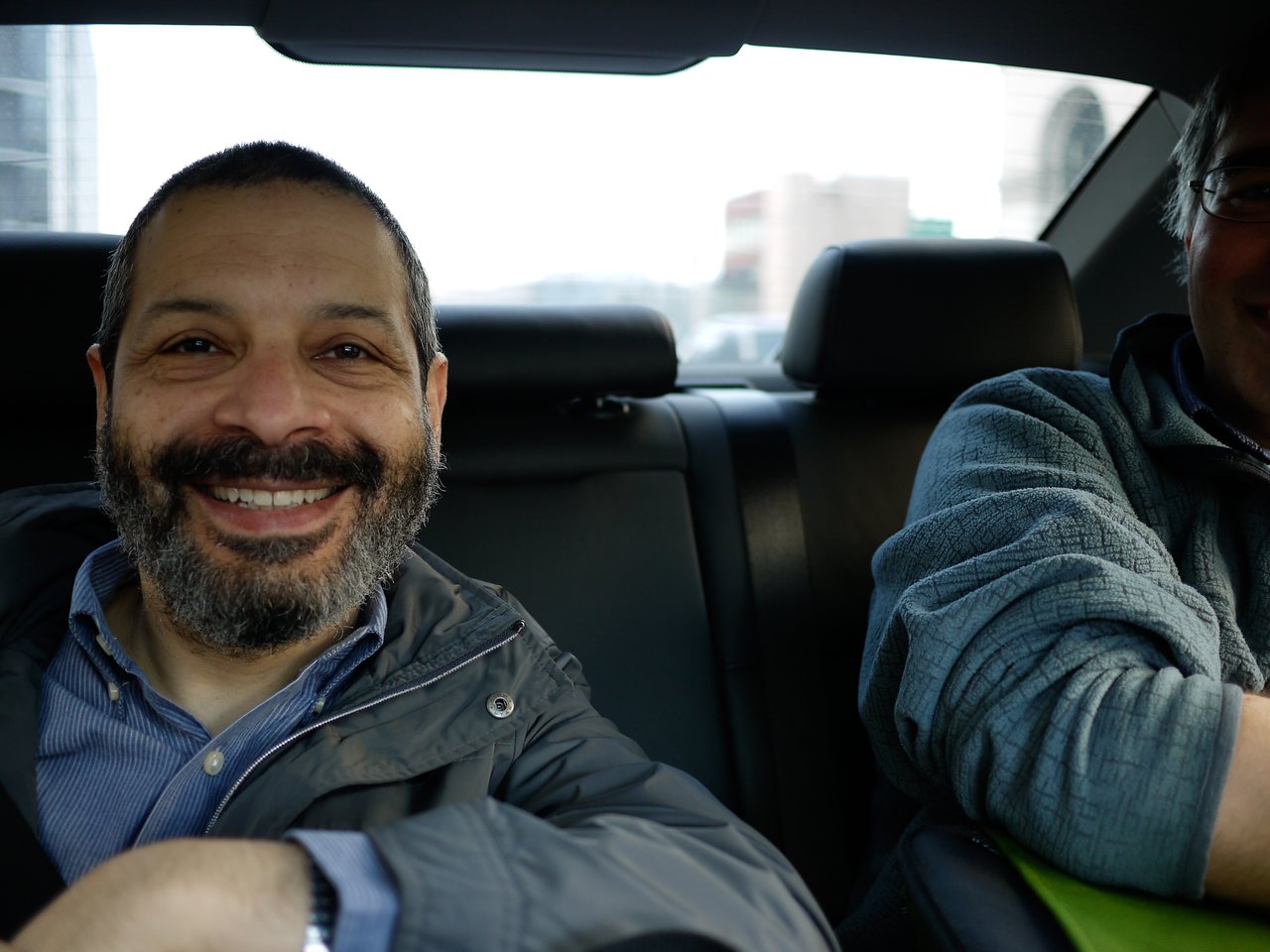 A smiling man with a beard sits in the backseat of a car, with another person partially visible.