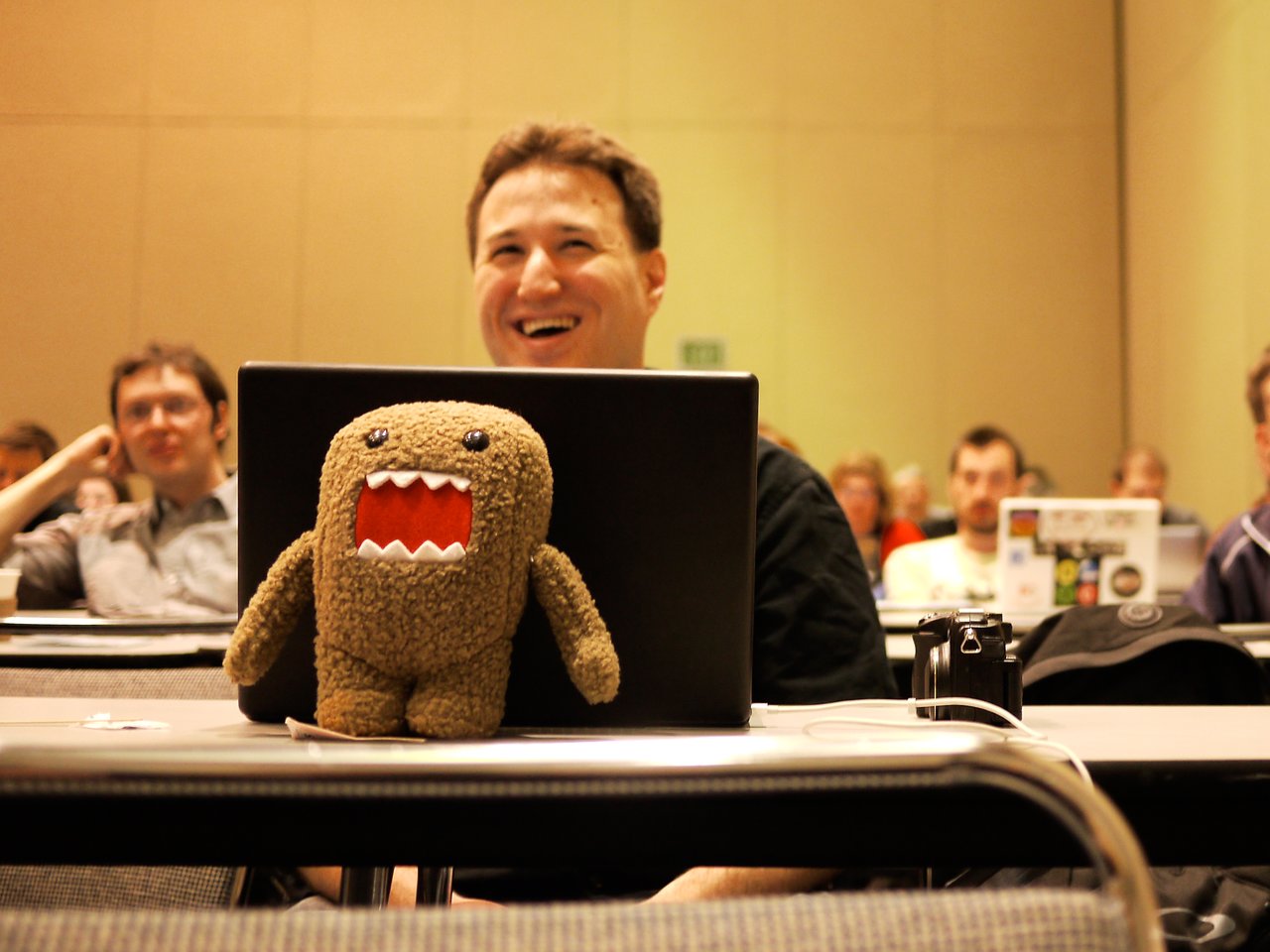 A small plush toy with an open mouth sits on a table in front of a laptop at a conference.