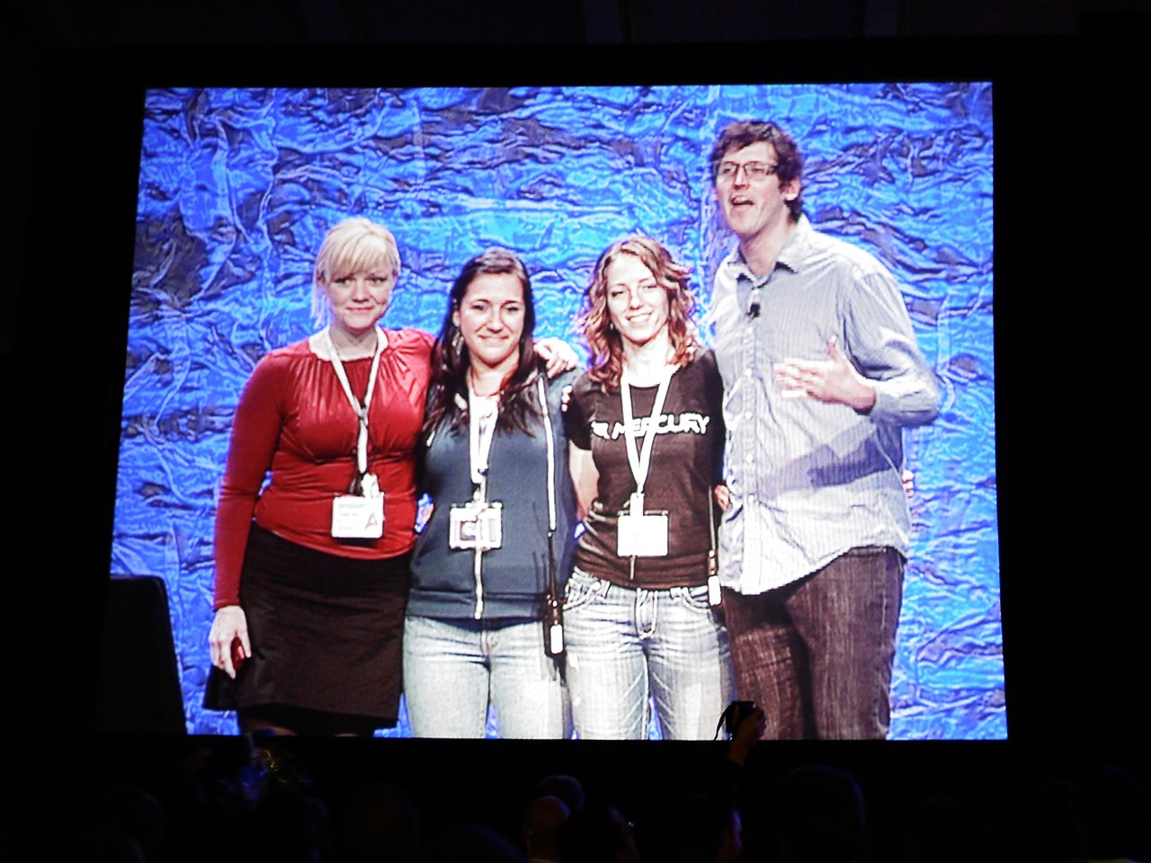 A projected image shows four DrupalCon San Francisco 2010 organizers standing together on stage during the closing session.
