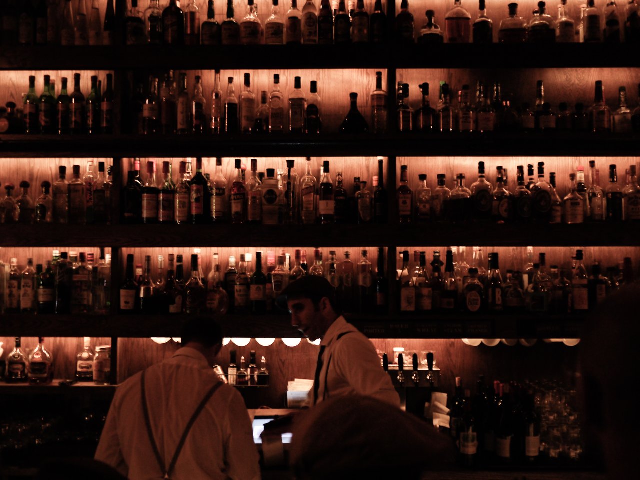 Bartenders in white shirts and suspenders serve drinks at a dimly lit bar with shelves of liquor bottles.