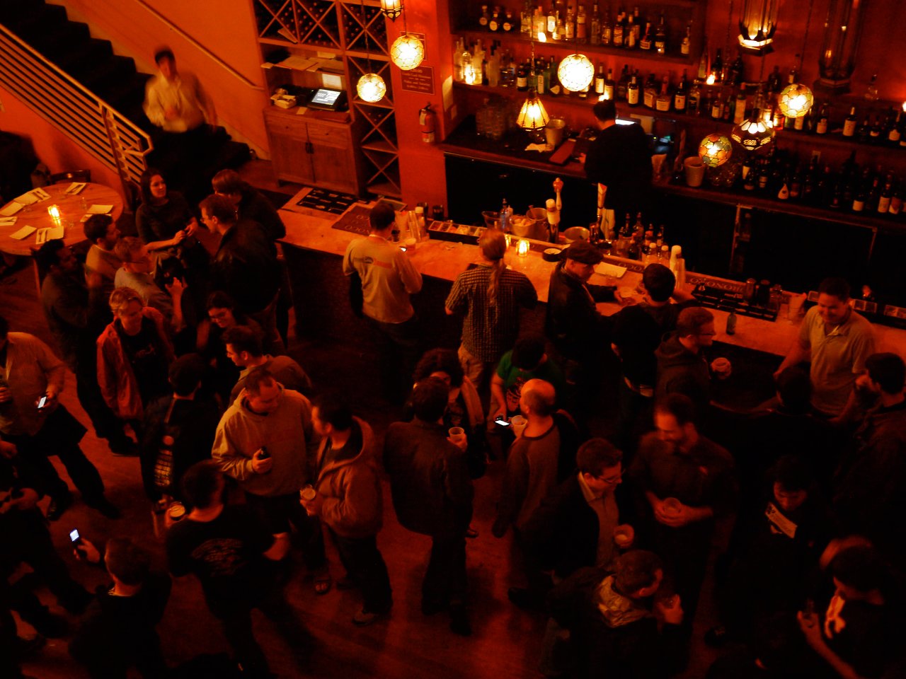A crowded bar with people socializing, holding drinks, and chatting while a bartender serves drinks behind the counter.