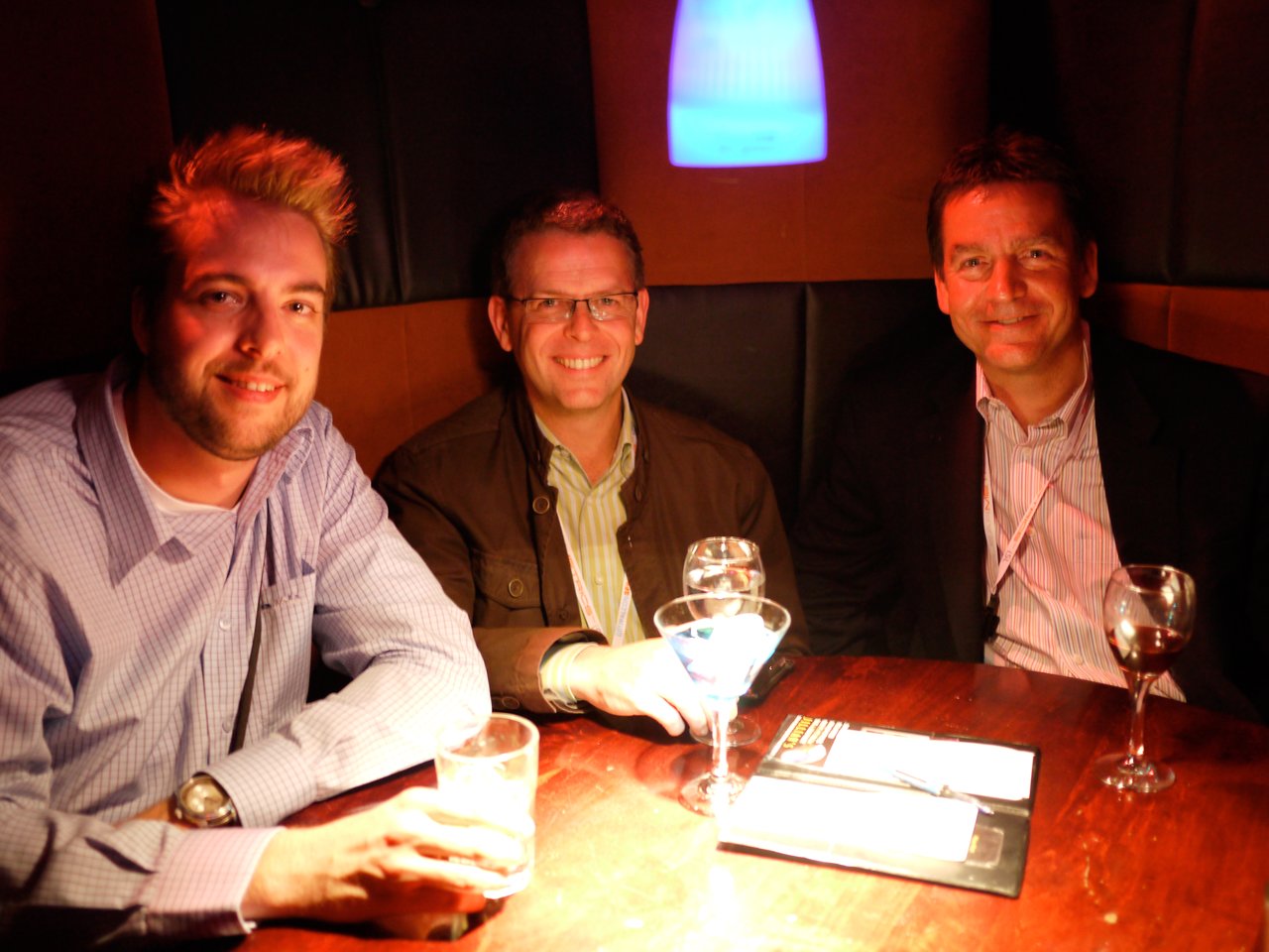 Three men sit at a dimly lit table, smiling and holding drinks at a social gathering.