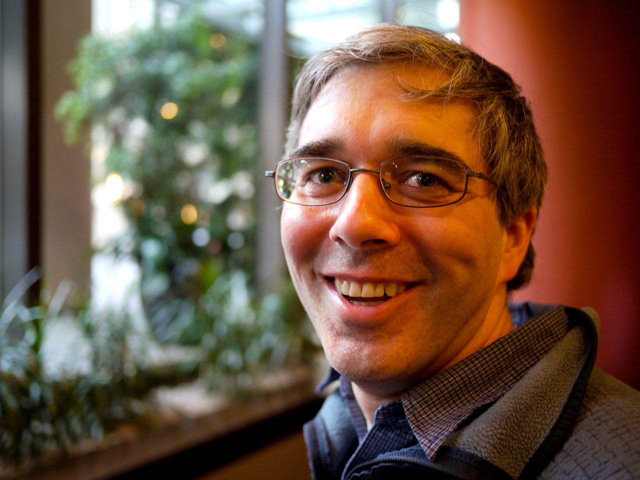 A man wearing glasses and a jacket smiles at the camera in an indoor setting with plants in the background.
