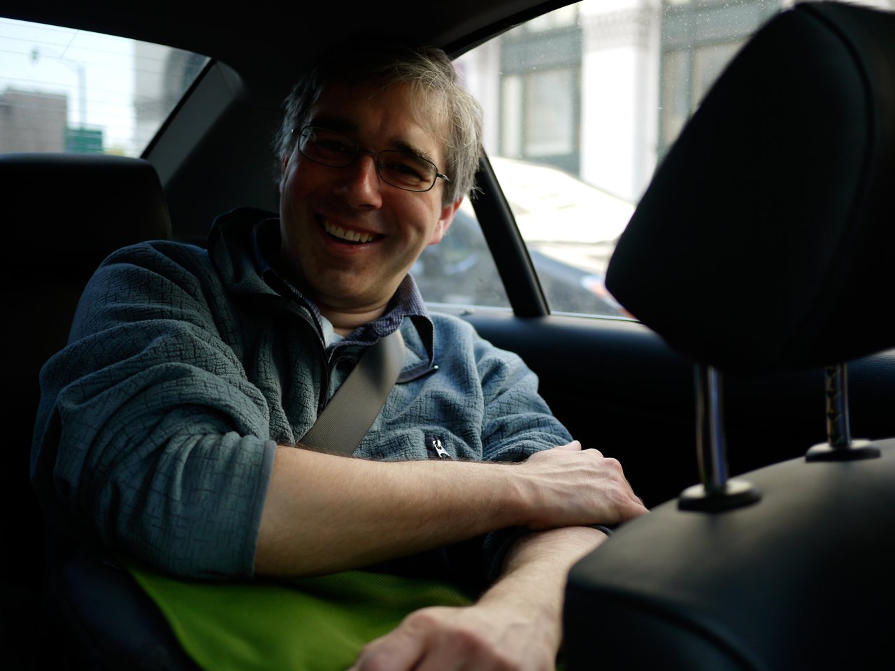 A man wearing glasses and a jacket smiles while sitting in the backseat of a car with a seatbelt on.