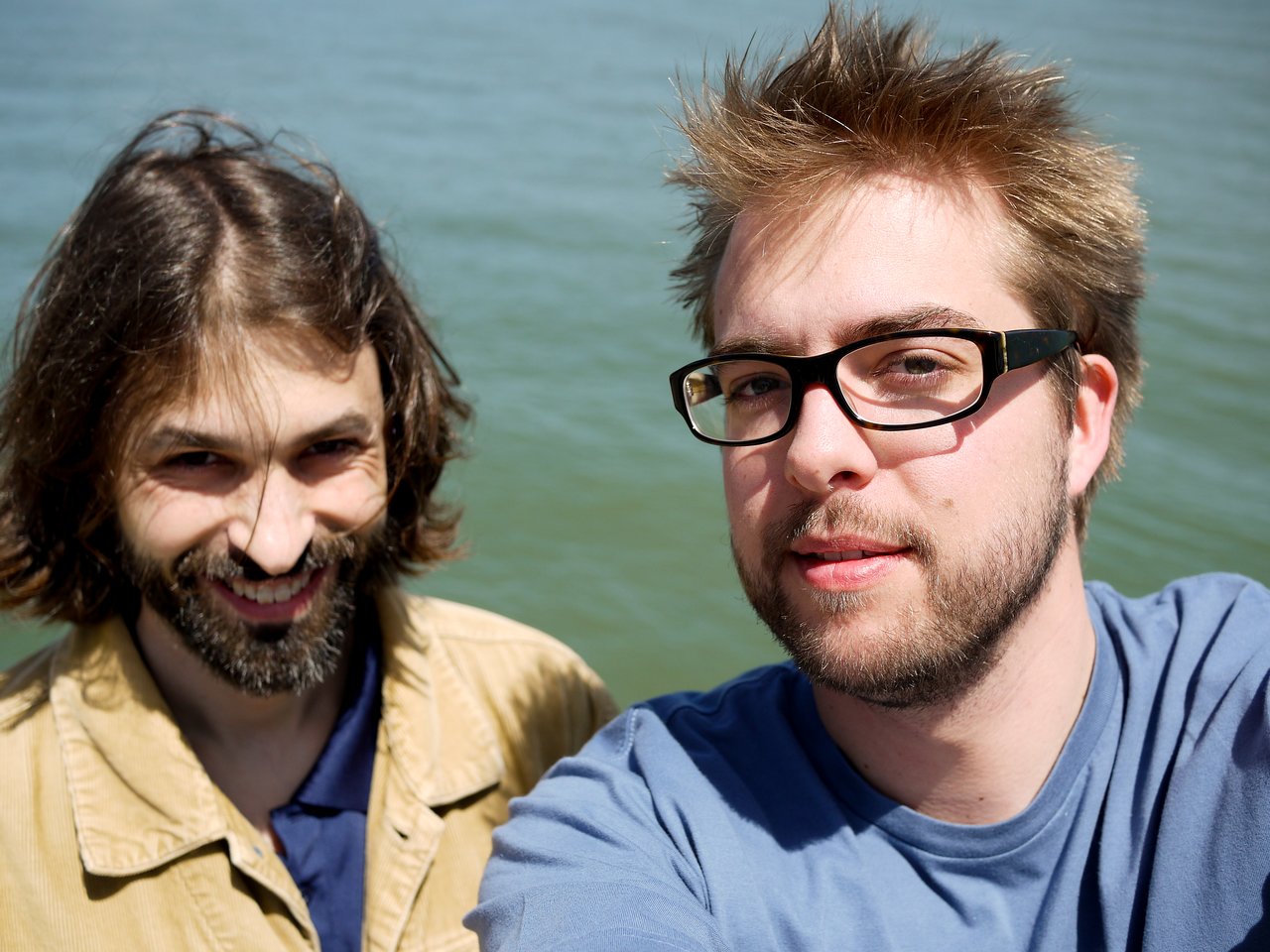 Two men standing outdoors near water, one taking a selfie while both smile at the camera.