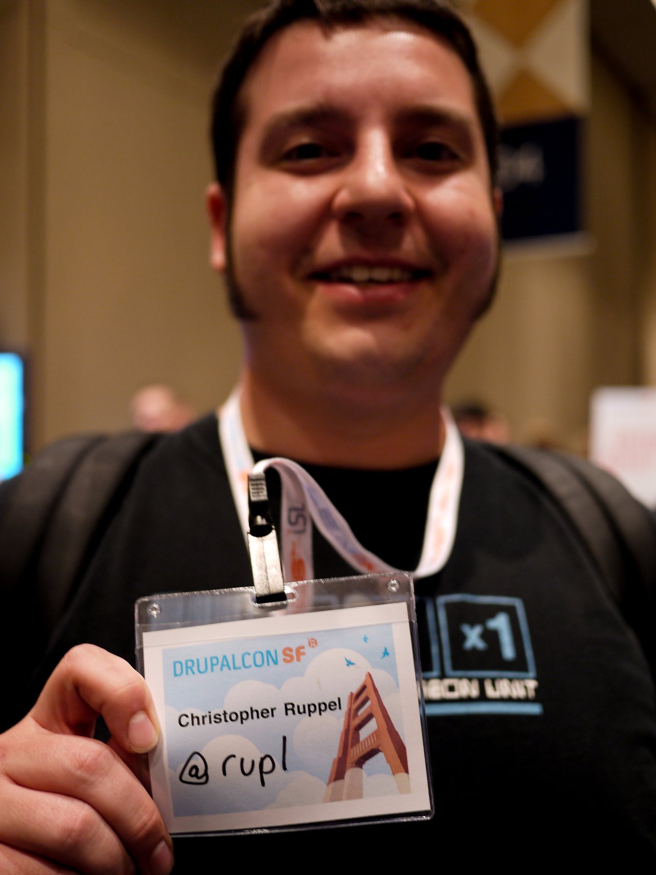 A person at DrupalCon San Francisco 2010 holds up a name badge that reads "Christopher Ruppel @rupl.