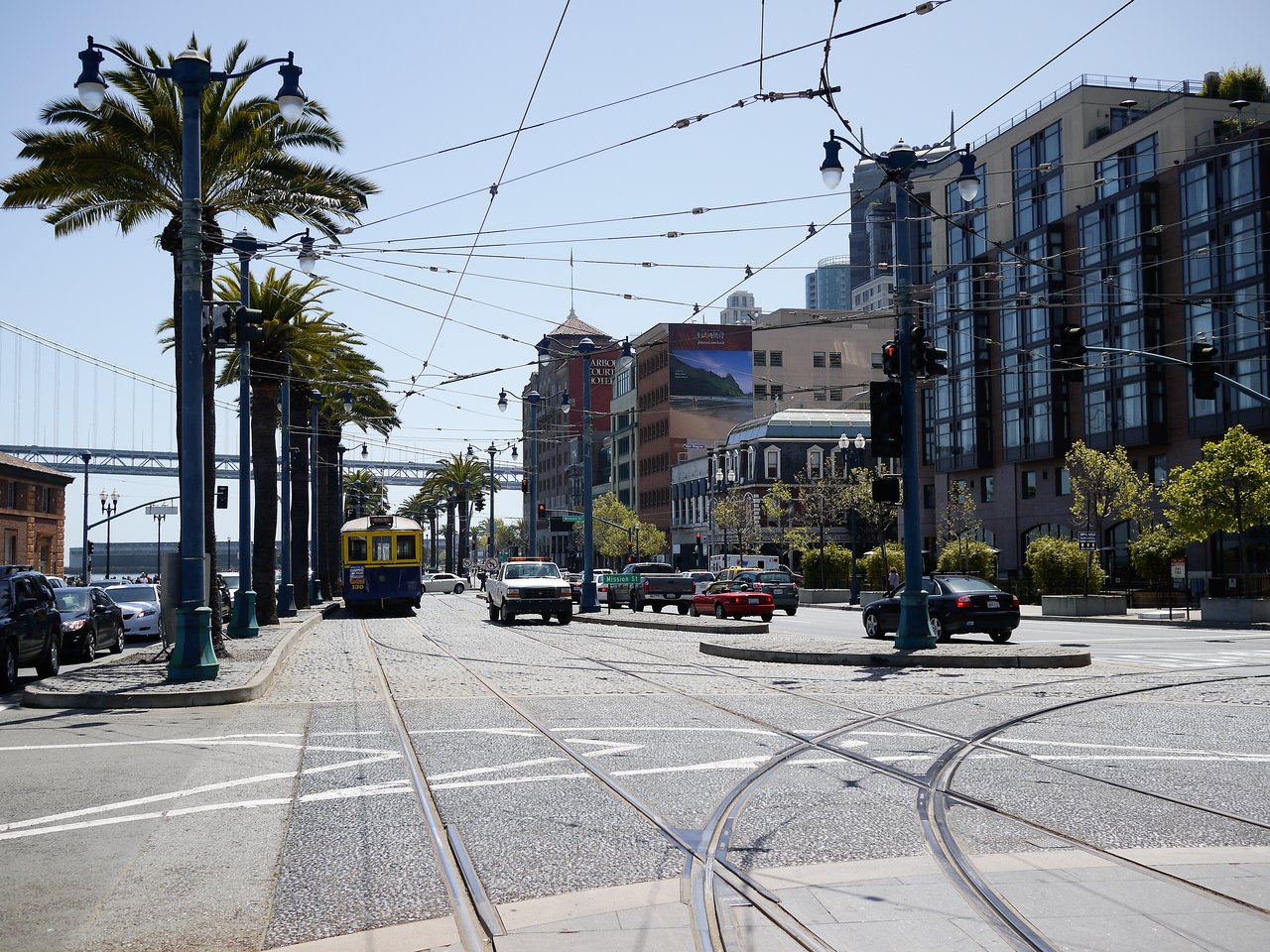 A street in San Francisco with a yellow and blue streetcar, cars, palm trees, and overhead tram wires.