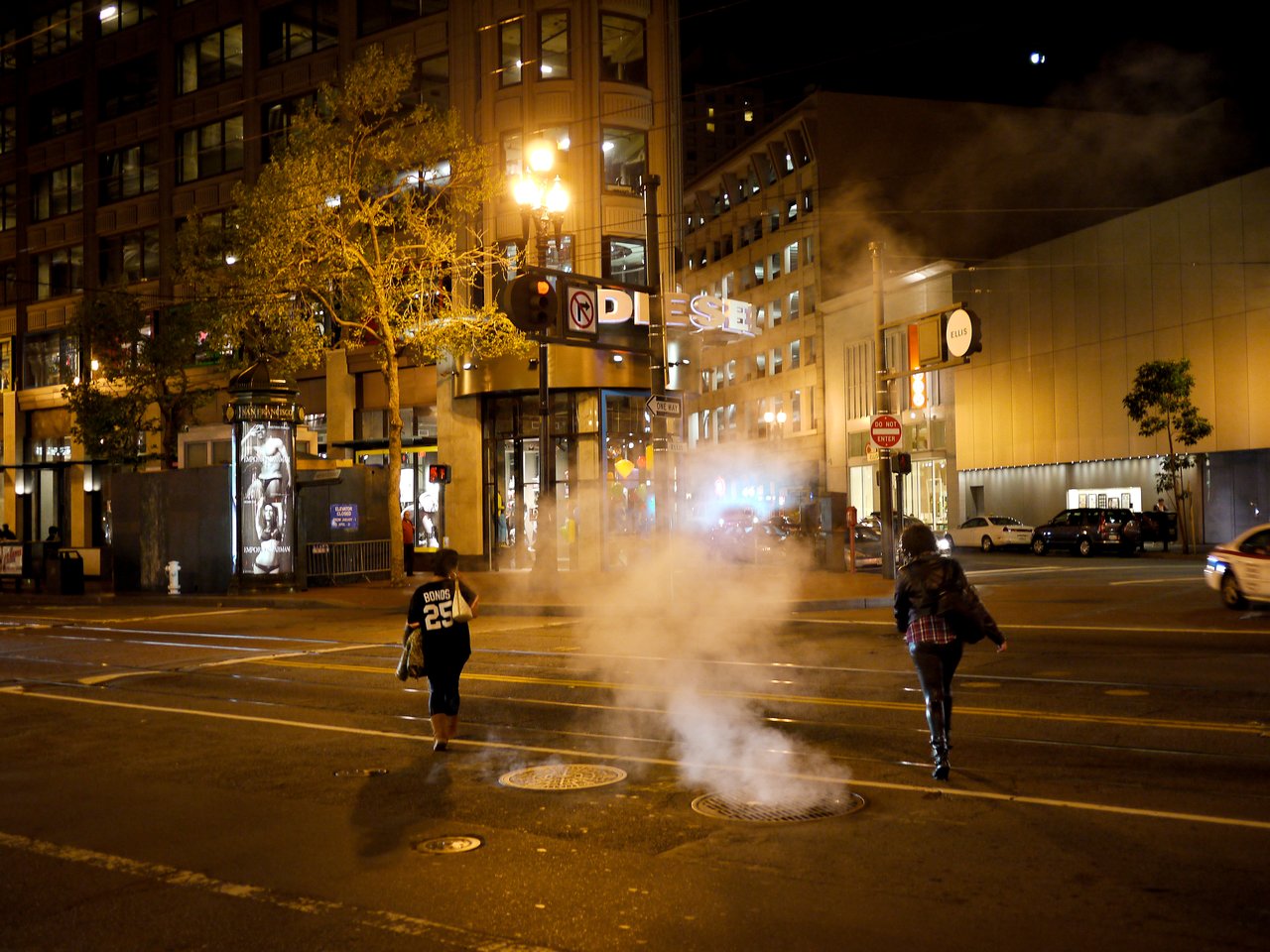 Two people walk across a city street at night, passing steam rising from a manhole.