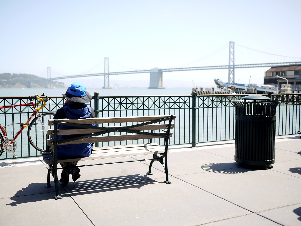 A person wearing a blue hat sits on a bench, looking out at the water and a large bridge.
