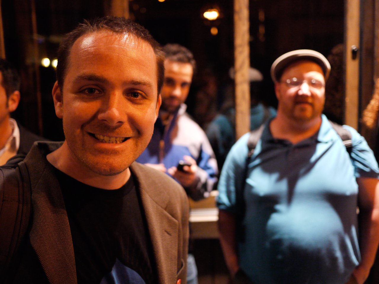 A man in the foreground smiles at the camera, while two other people stand slightly out of focus behind him.