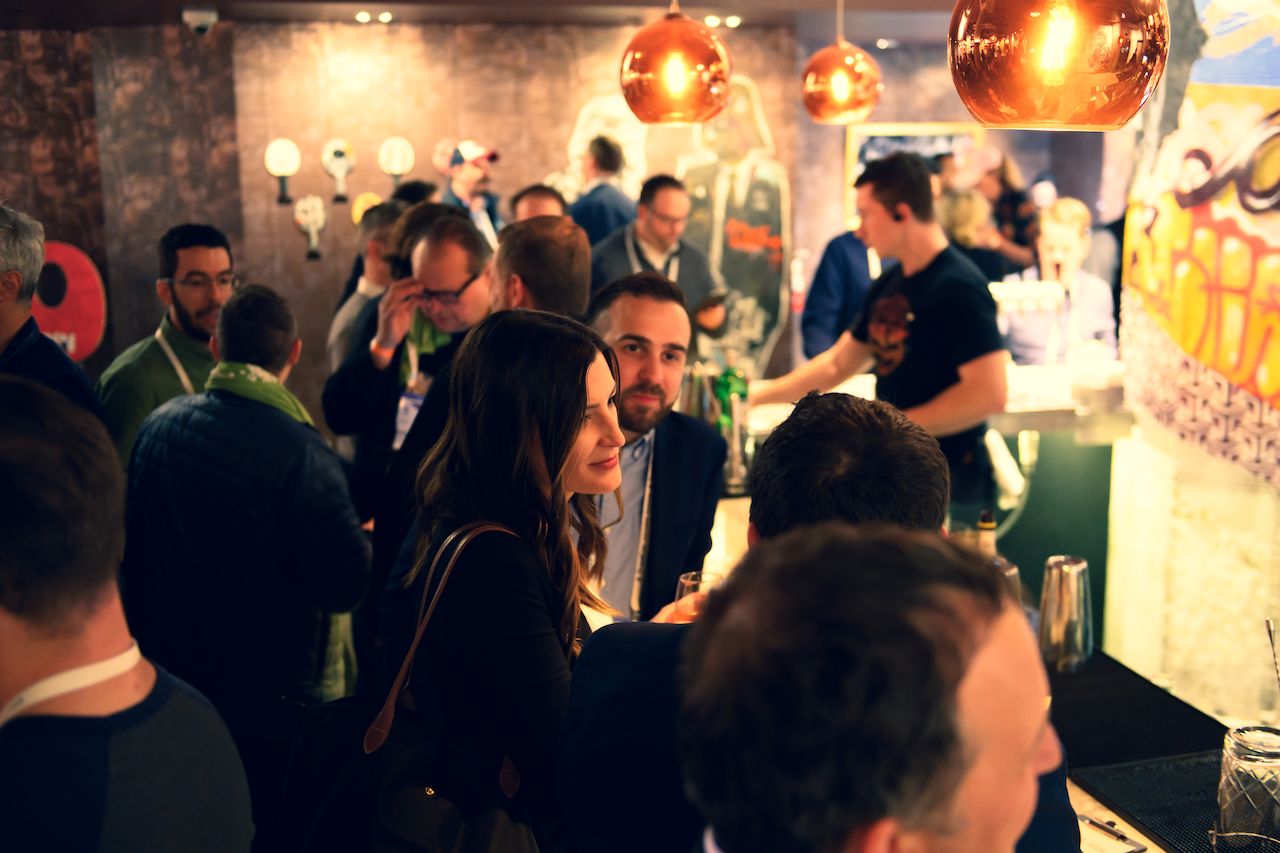 A woman with long hair smiles while talking to a man in a crowded bar or networking event.
