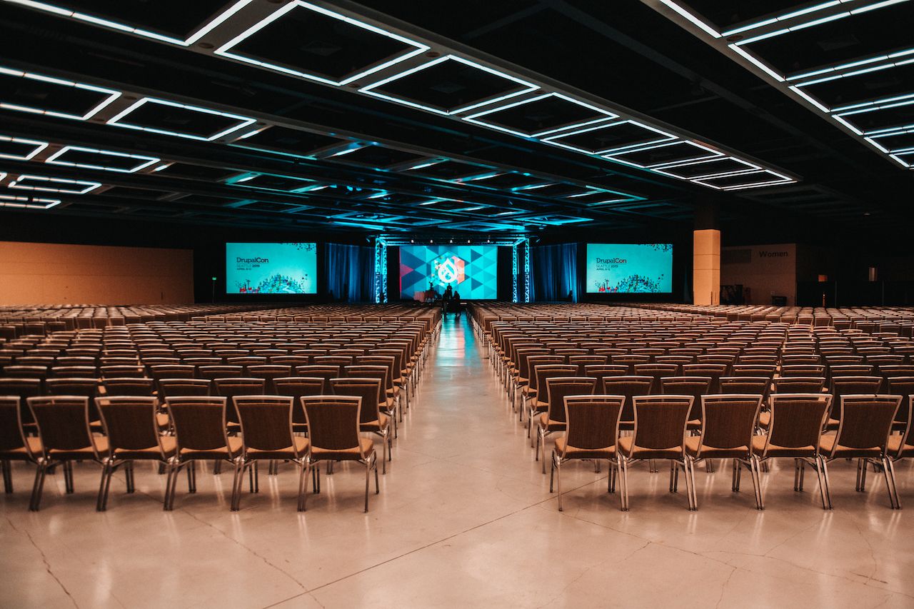Large conference hall with rows of empty chairs facing a stage with big screens showing "DrupalCon" branding.