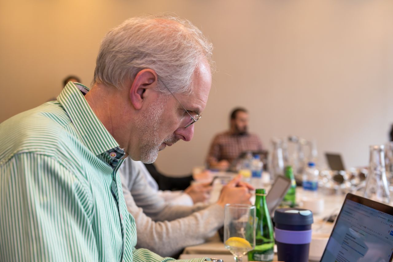 Man in a green striped shirt focuses on his laptop during a meeting with others working in the background.