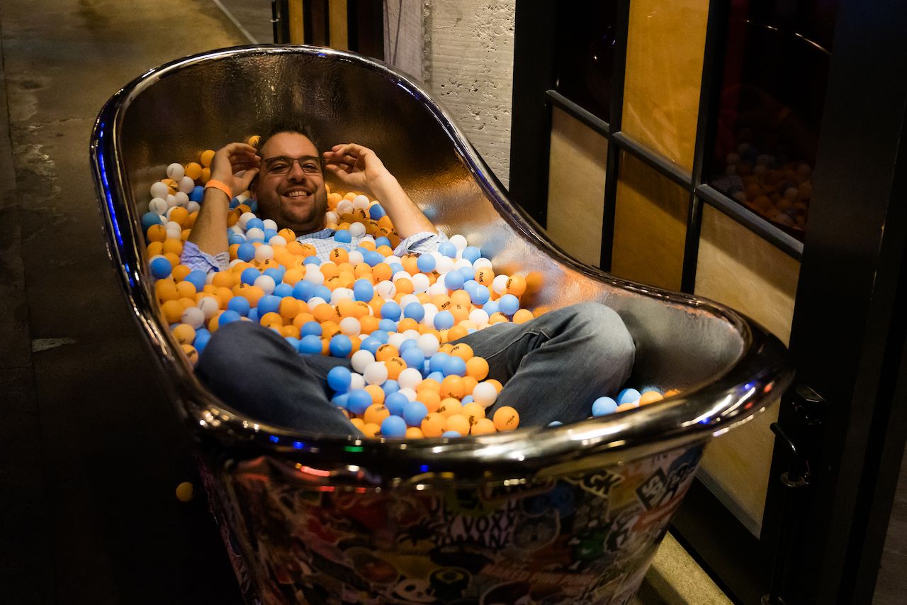 A man smiles while lying in a bathtub filled with colorful ping pong balls.