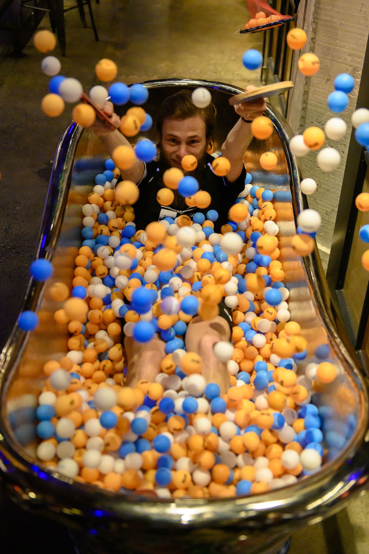 A person sits in a bathtub filled with colorful ping pong balls, tossing them in the air while smiling.