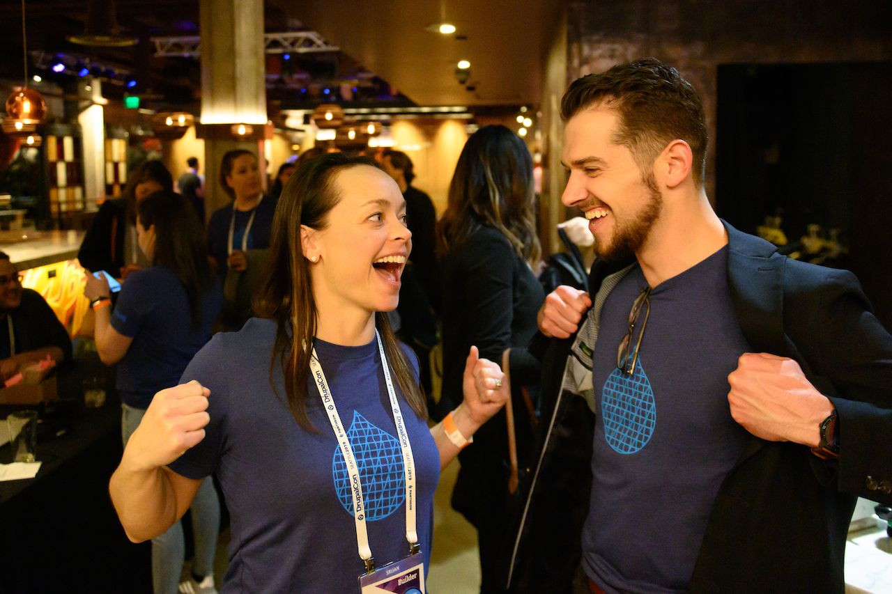 Two people smile and gesture excitedly, showing matching Acquia t-shirts at a social event.
