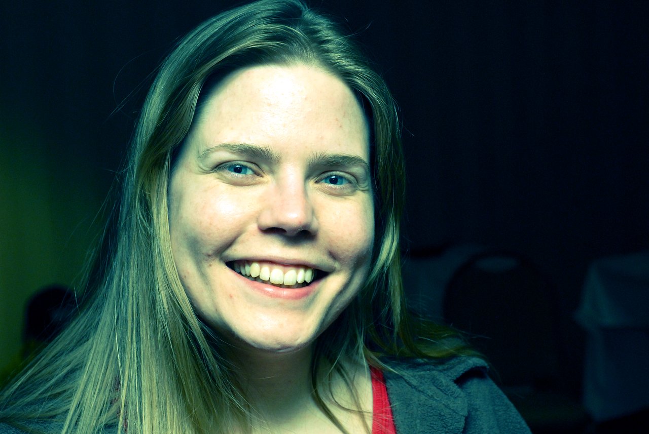 A person with long hair smiles at the camera in a dimly lit indoor setting.