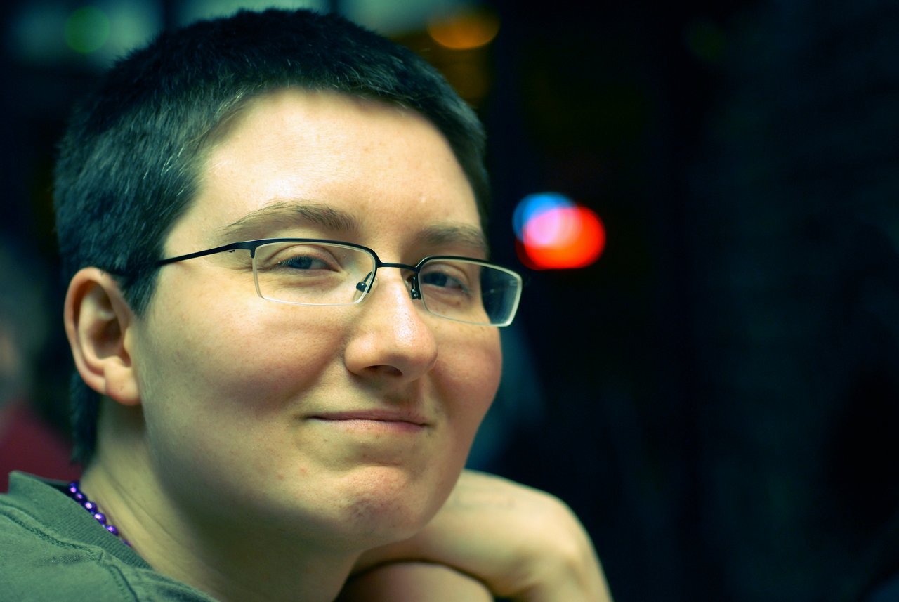 A person with short hair and glasses smiles slightly while resting their chin on their hand in a dimly lit setting.