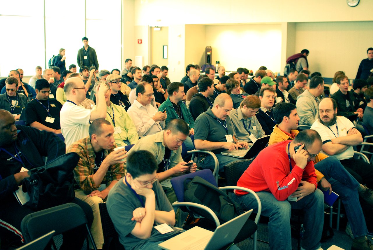 A large group of conference attendees sit closely together, using laptops, phones, and conversing during a session.