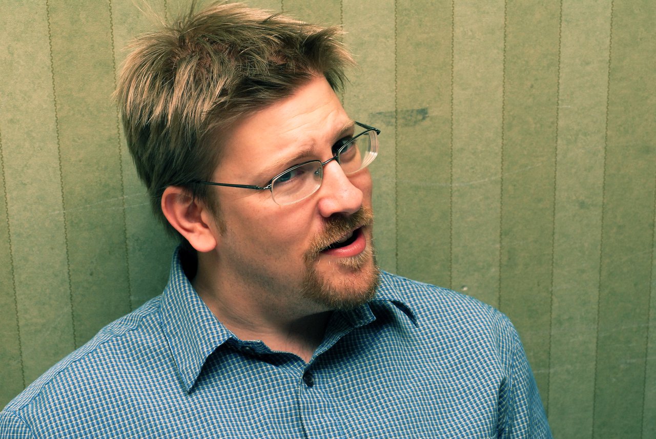A man wearing glasses and a checkered shirt speaks while looking slightly to the side against a green background.