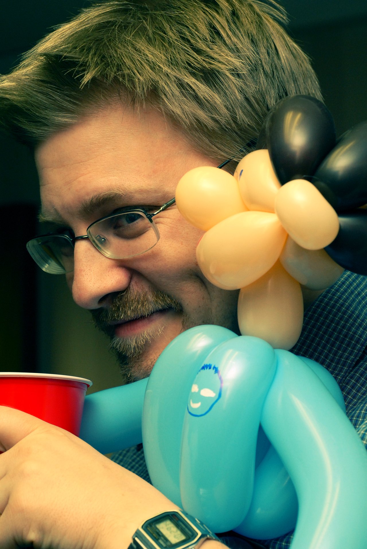 A person with glasses smiles while holding a red cup, with a balloon figure resting on their shoulder.