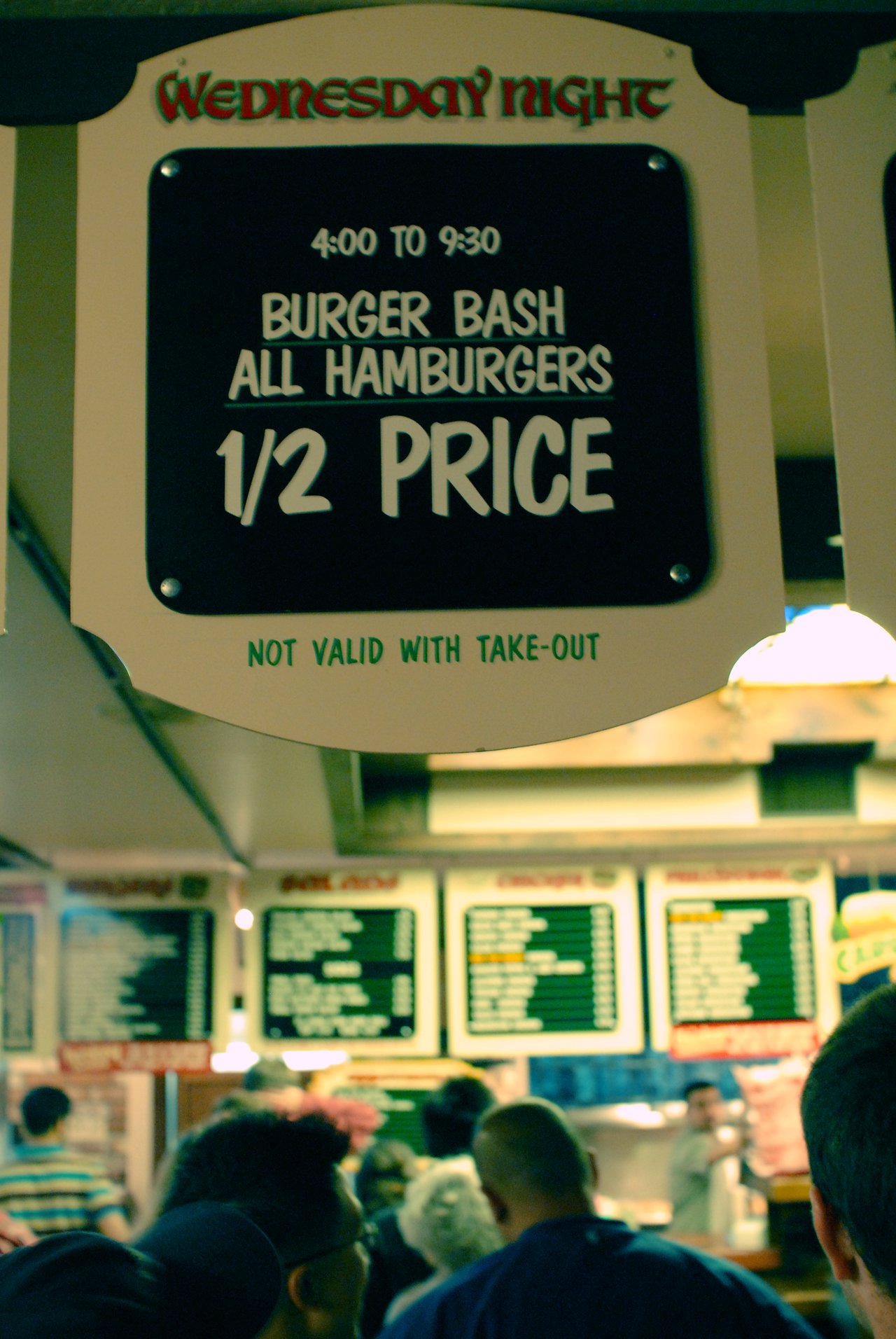 A crowd waits in line at a burger restaurant under a sign advertising half-price hamburgers for a special event.