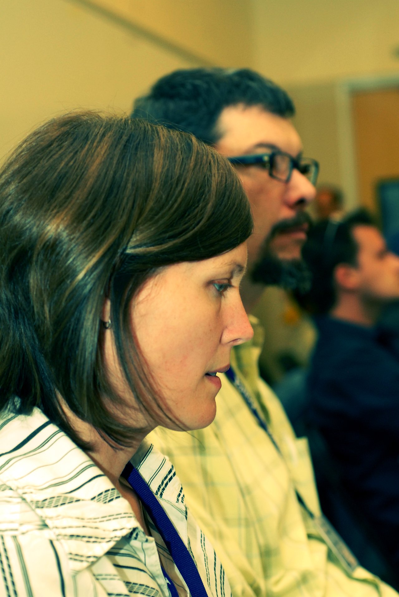 A woman with short brown hair attentively listens at a conference, with two men seated behind her.
