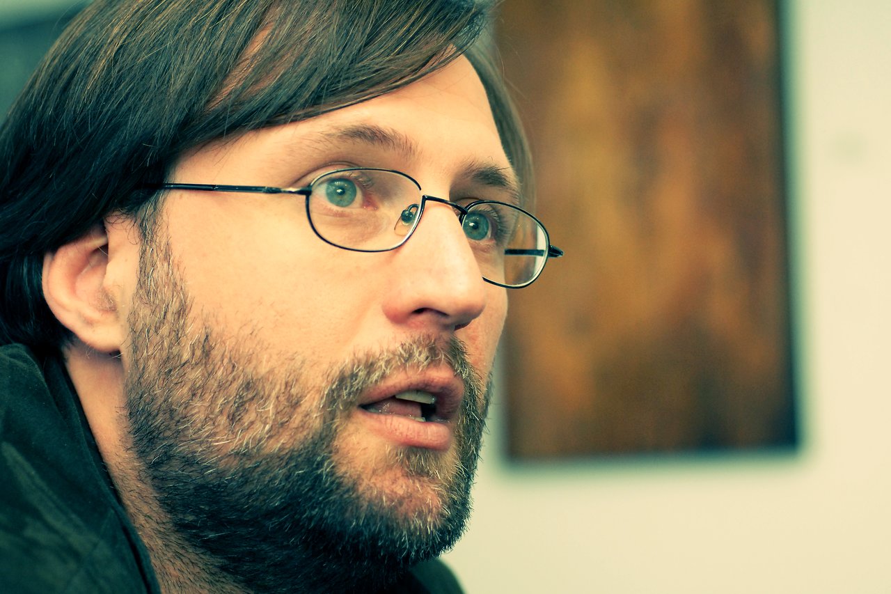 A man with glasses and a beard looks intently off-camera, appearing engaged in conversation or deep thought.