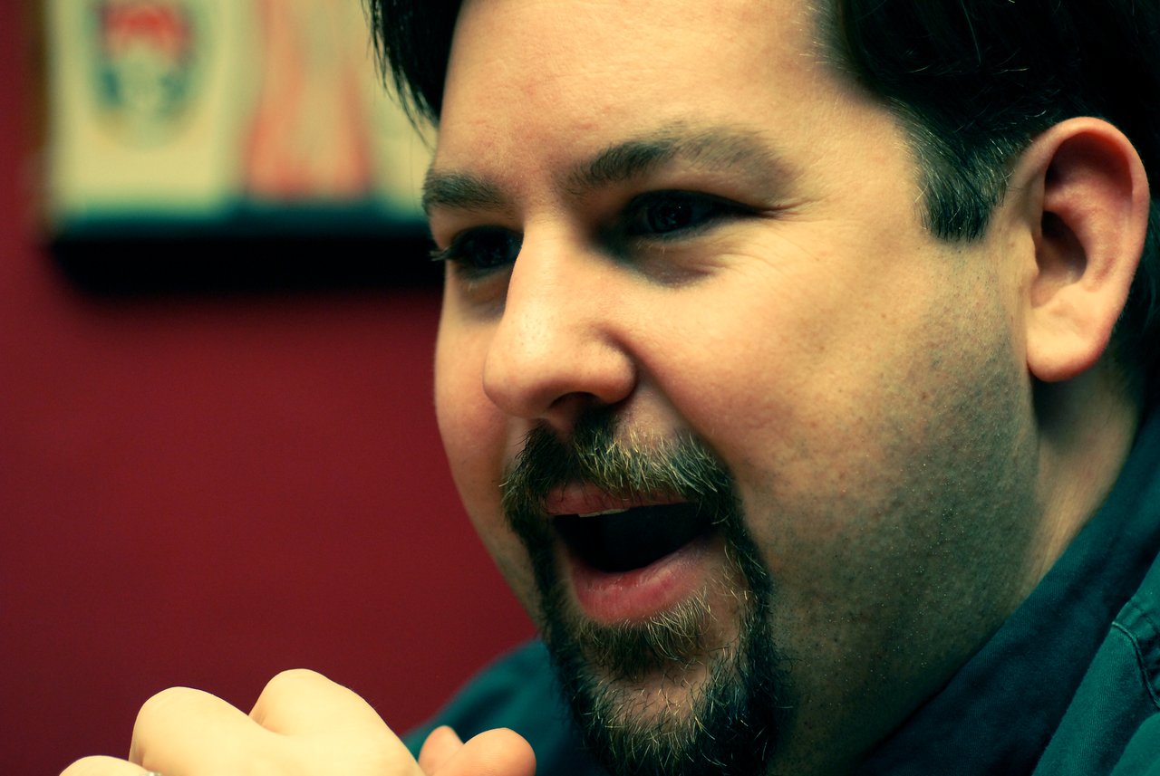 A man with a beard and mustache is speaking with an engaged expression, hands clasped near his face.