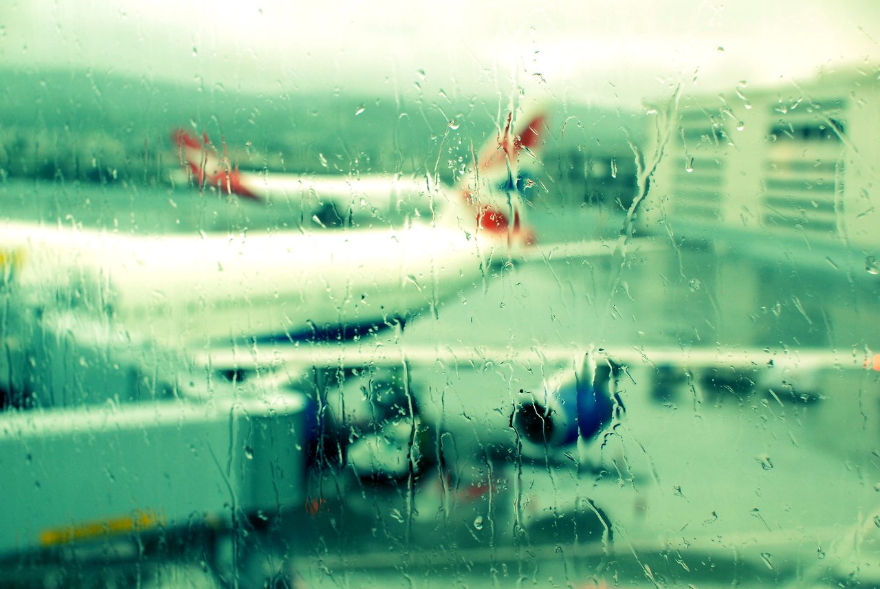 Rain-covered airport window with a blurry view of airplanes and ground crew preparing for departure.