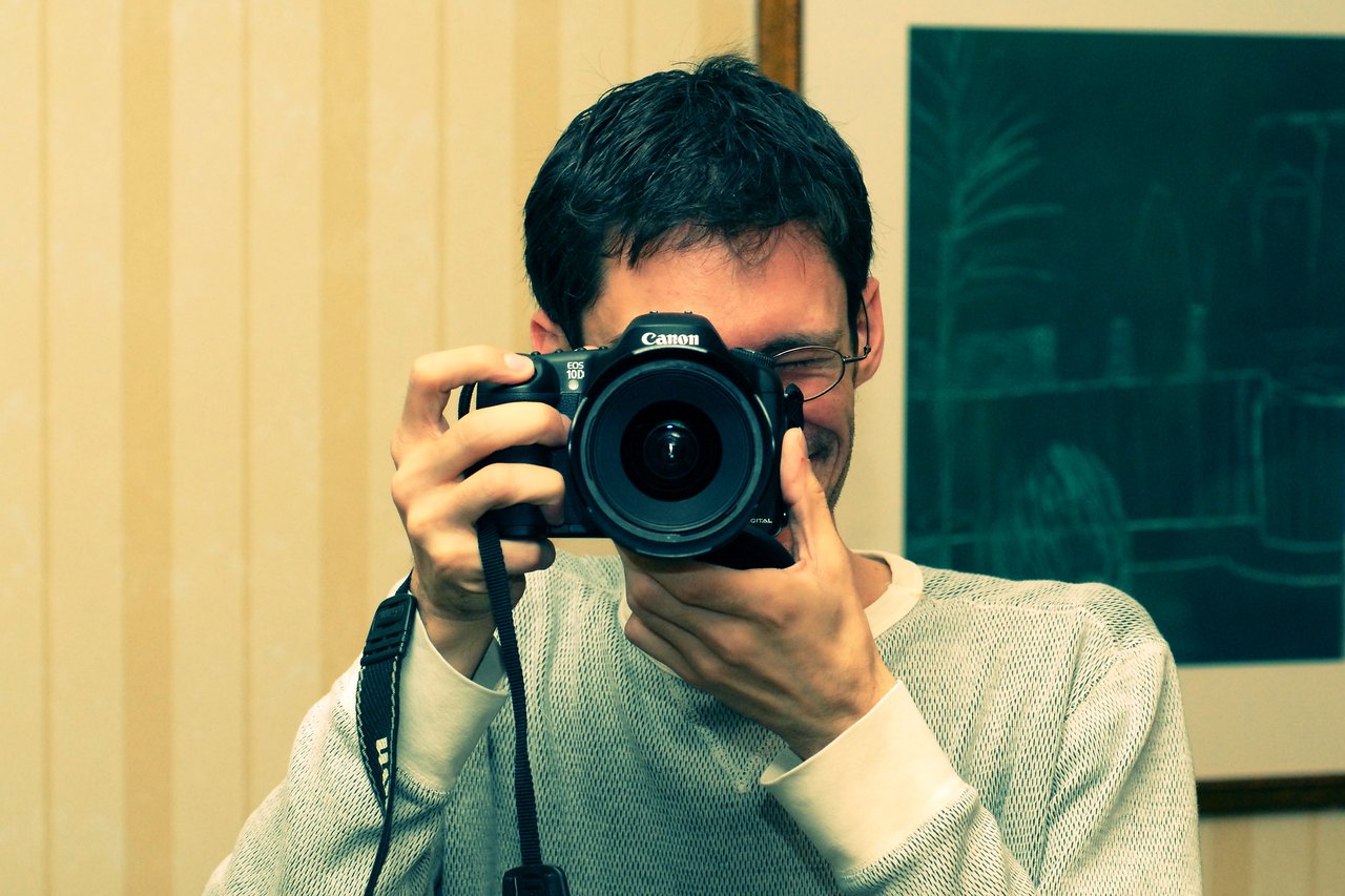 A person in a light-colored sweater holds a Canon EOS 10D camera, aiming it directly at the viewer.